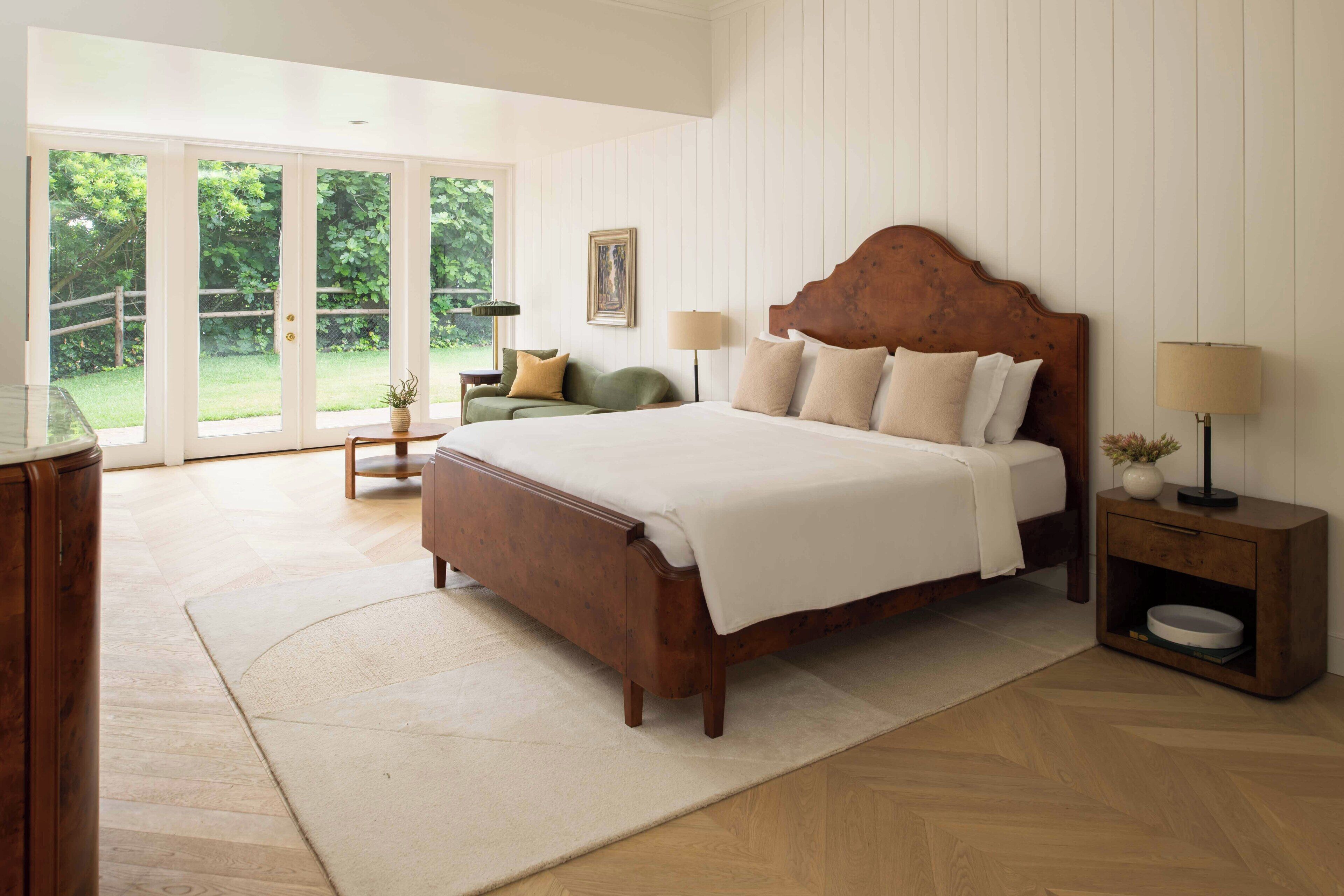 A bedroom with a large bed, a nightstand, a lamp, a chair, and a view of a garden through a sliding door.