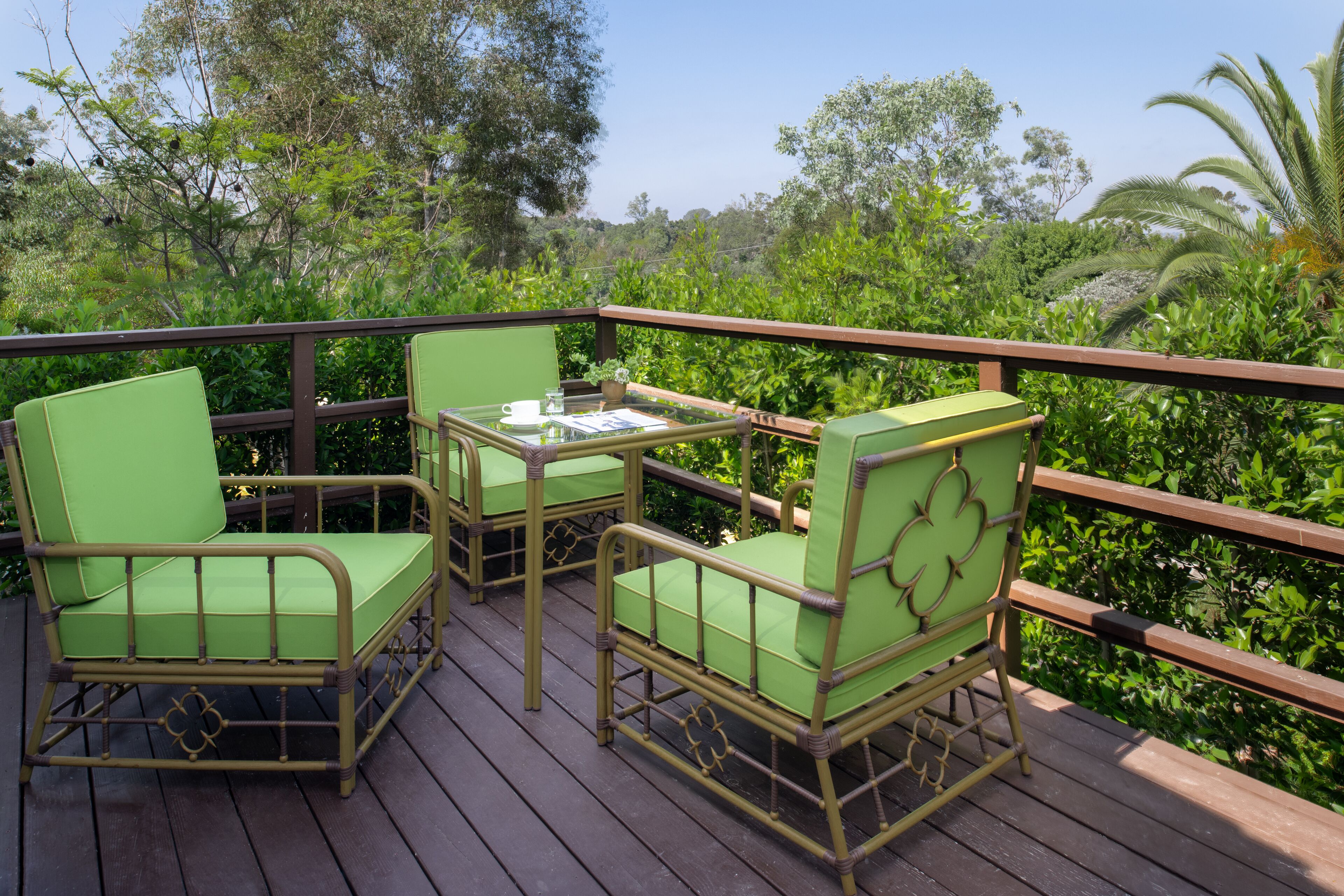 A wooden deck with green cushioned chairs and a small table, surrounded by lush greenery.