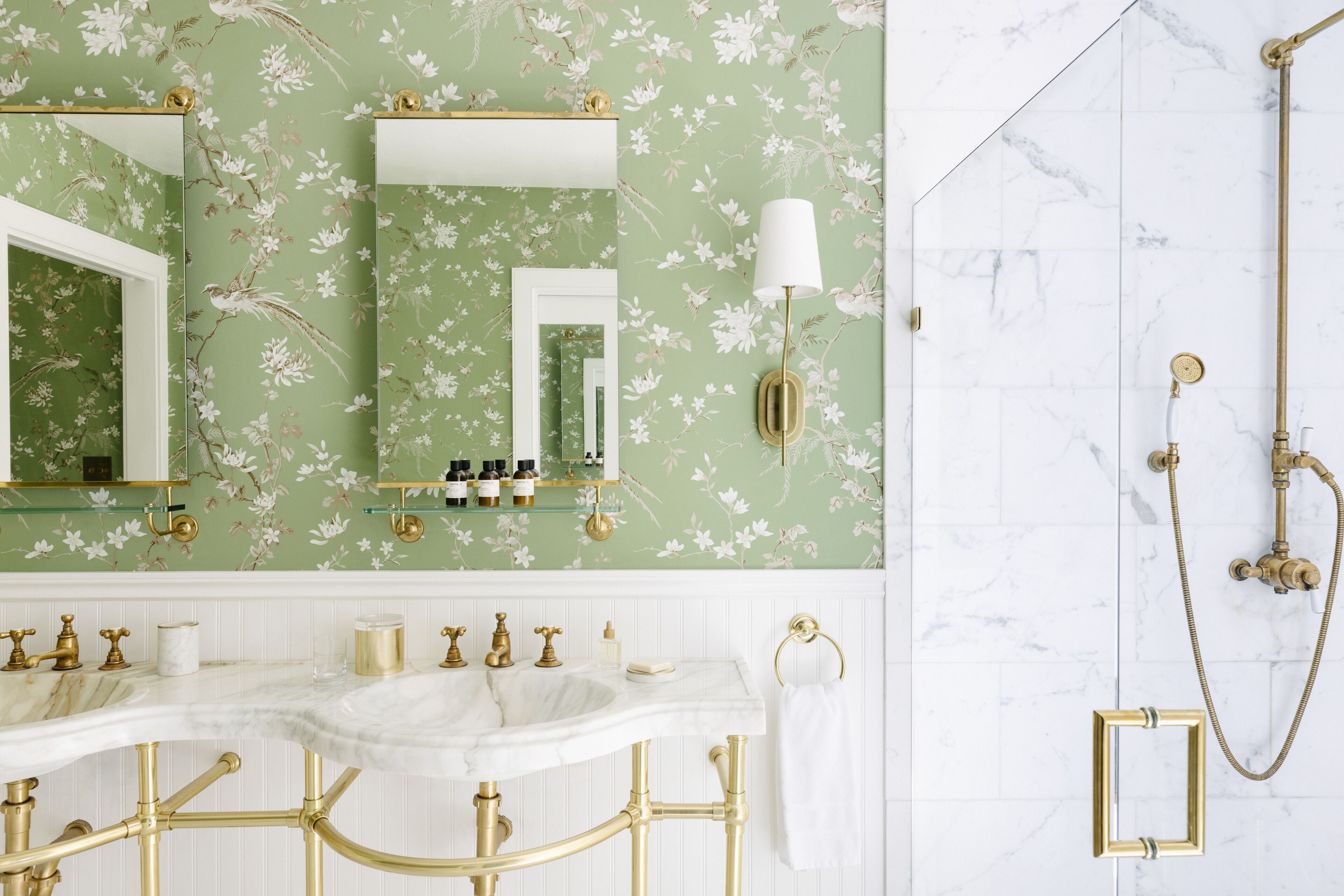 Un baño con papel tapiz floral, un lavamanos de mármol blanco, accesorios dorados y una ducha dorada.