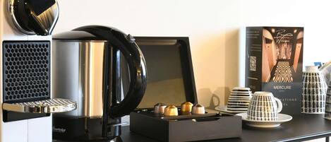 A coffee station with a coffee maker, a kettle, and a tray of pastries. There is a framed welcome sign and a stack of plates.
