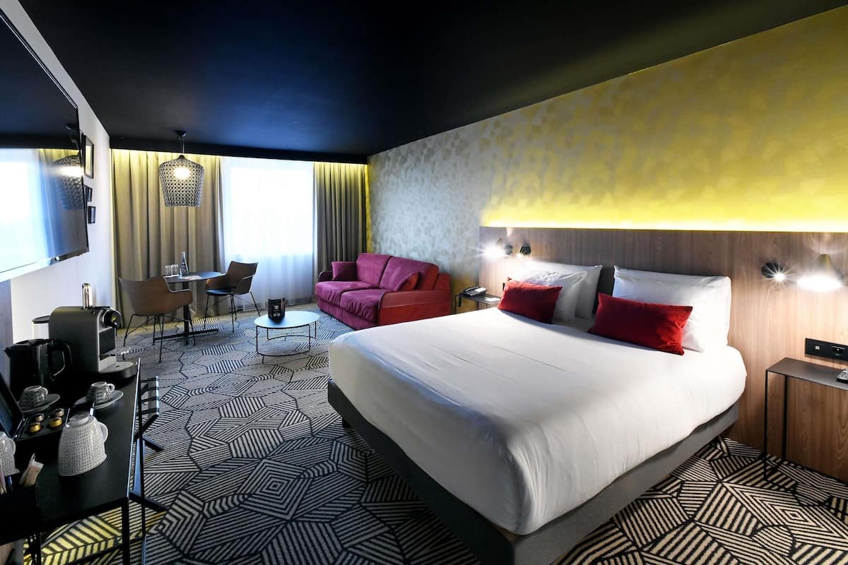 A hotel room with a large bed, a desk with a chair, a sofa, and a TV mounted on the wall.