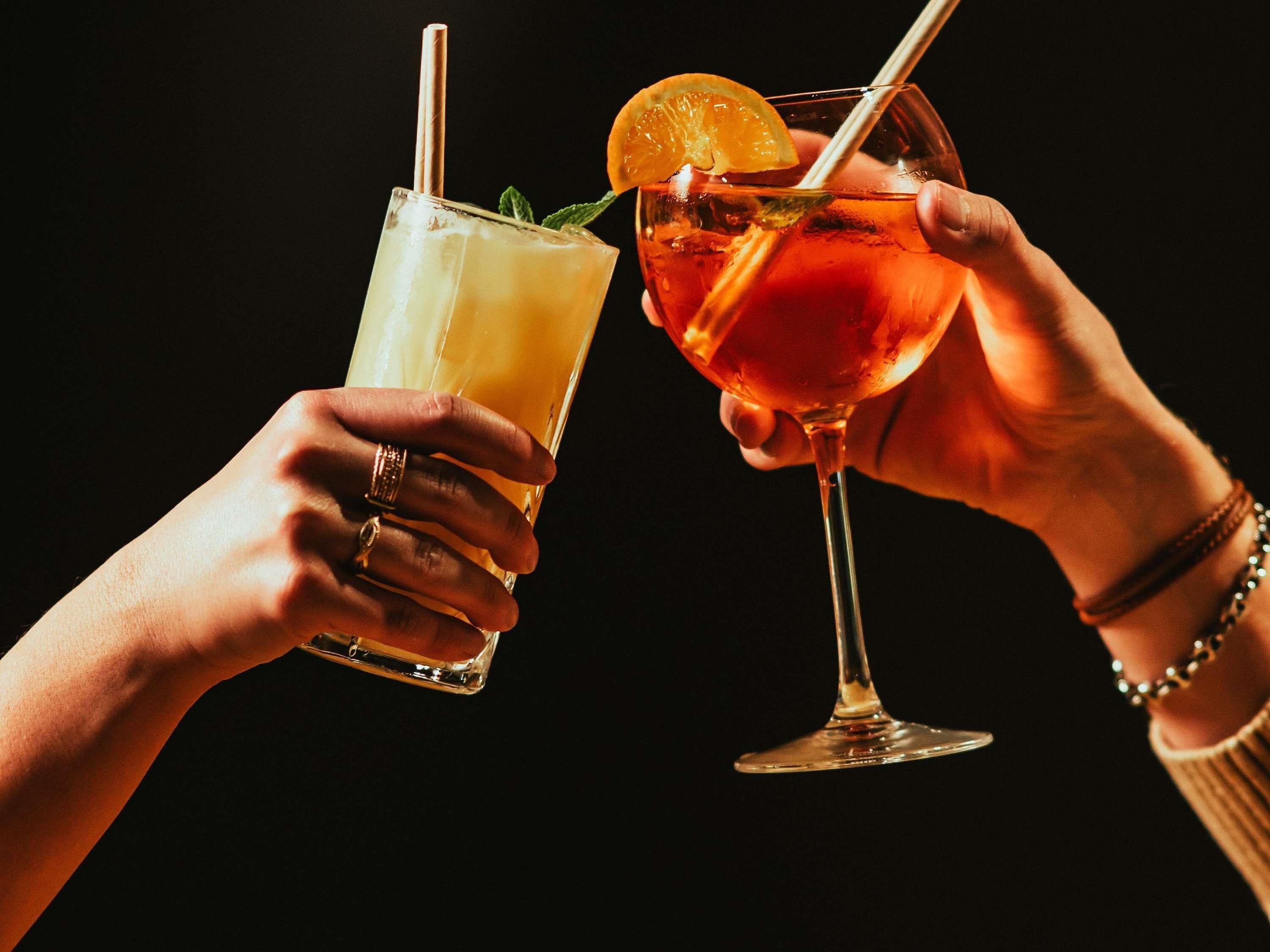 Zwei Hände halten Gläser mit Getränken, eines mit Limettenscheibe und Minze, das andere mit Orangenscheibe.