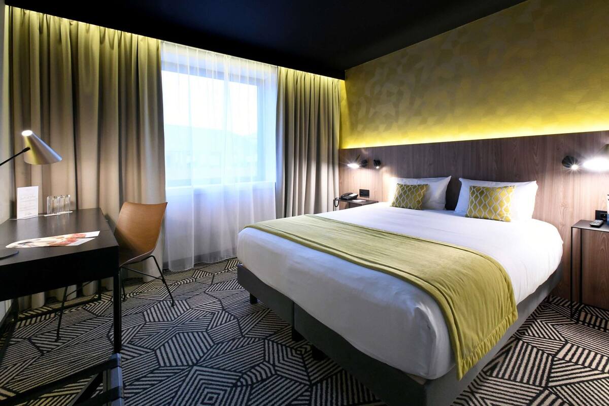 A hotel room with a large bed, a desk with a lamp, a chair, and a window with curtains.