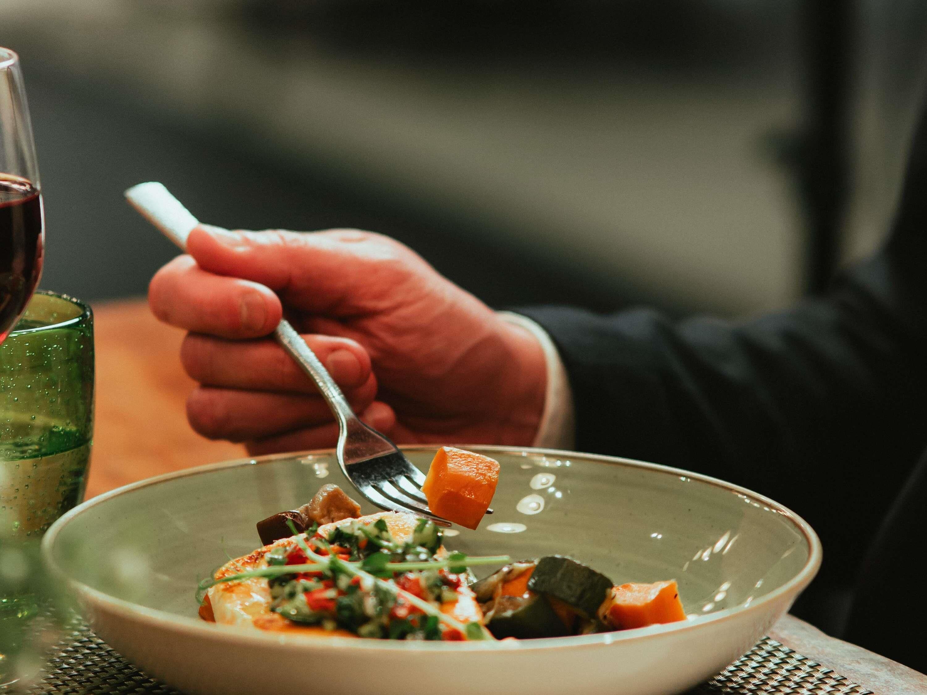 Una persona sosteniendo un tenedor sobre un bol de comida con verduras y un trozo de carne.