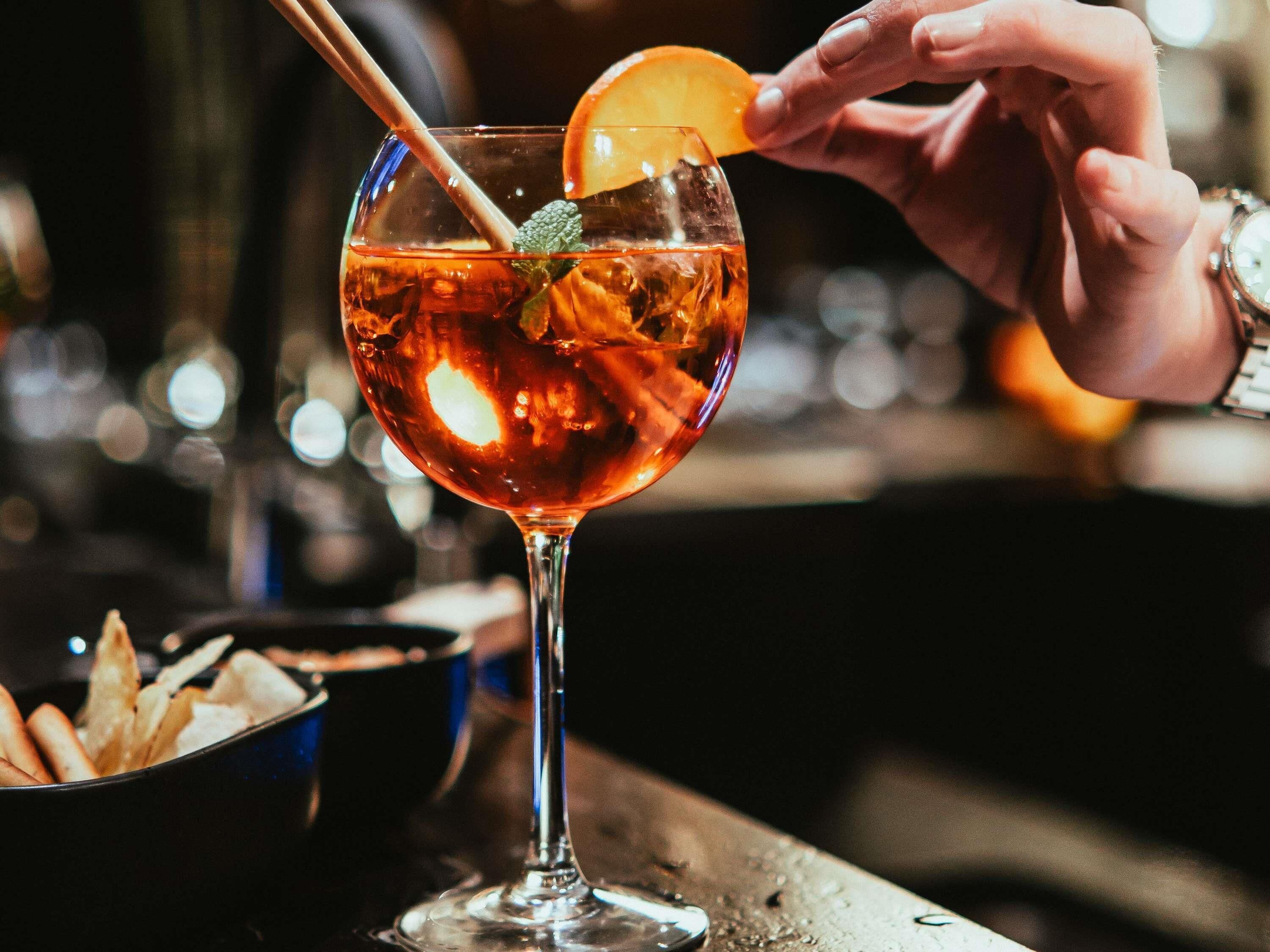 A cocktail glass with a slice of orange and mint leaves, a straw, and ice cubes.
