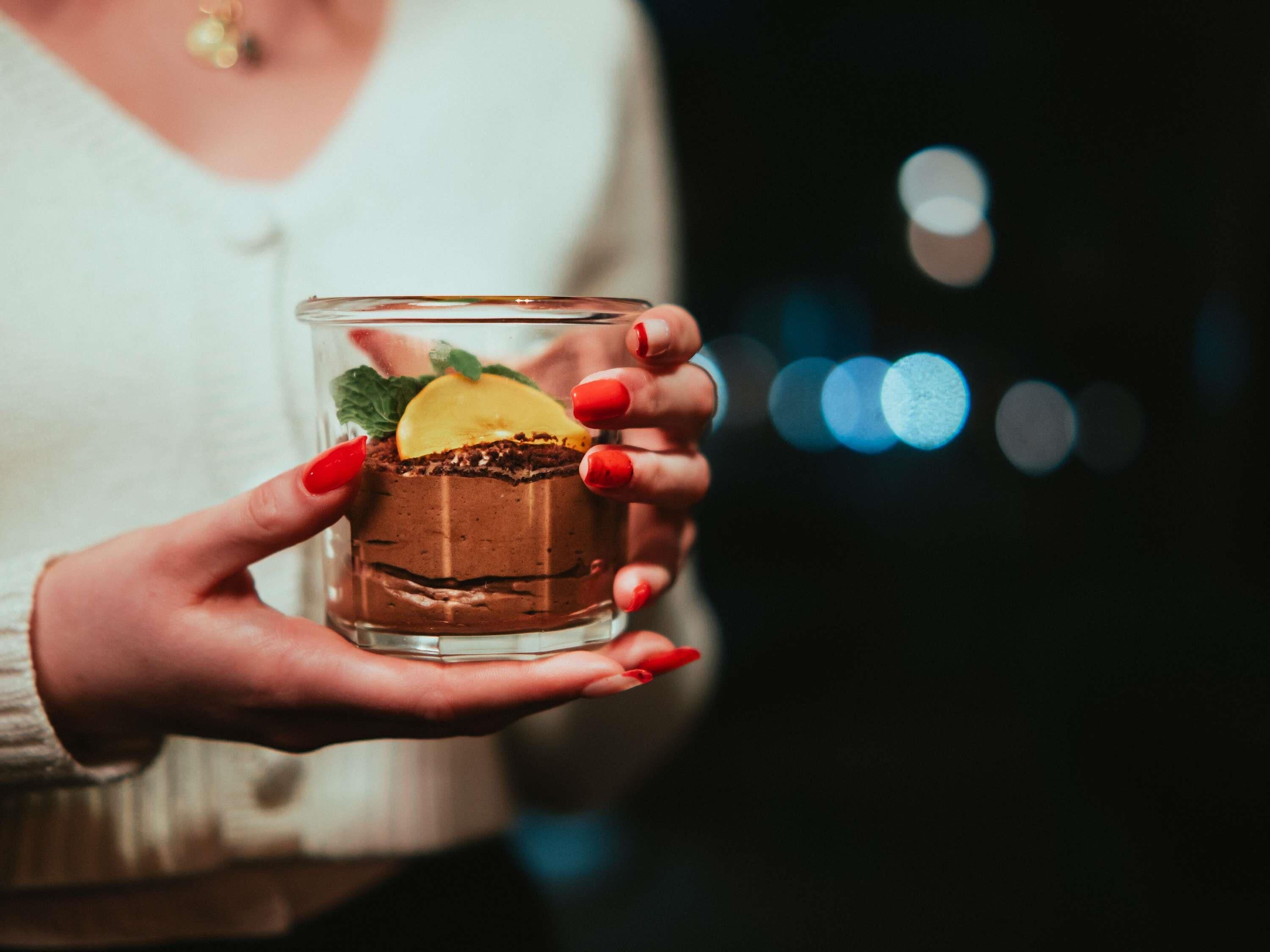 A person holding a glass with a layered dessert, possibly a cake or mousse, garnished with a slice of fruit and mint leaves.