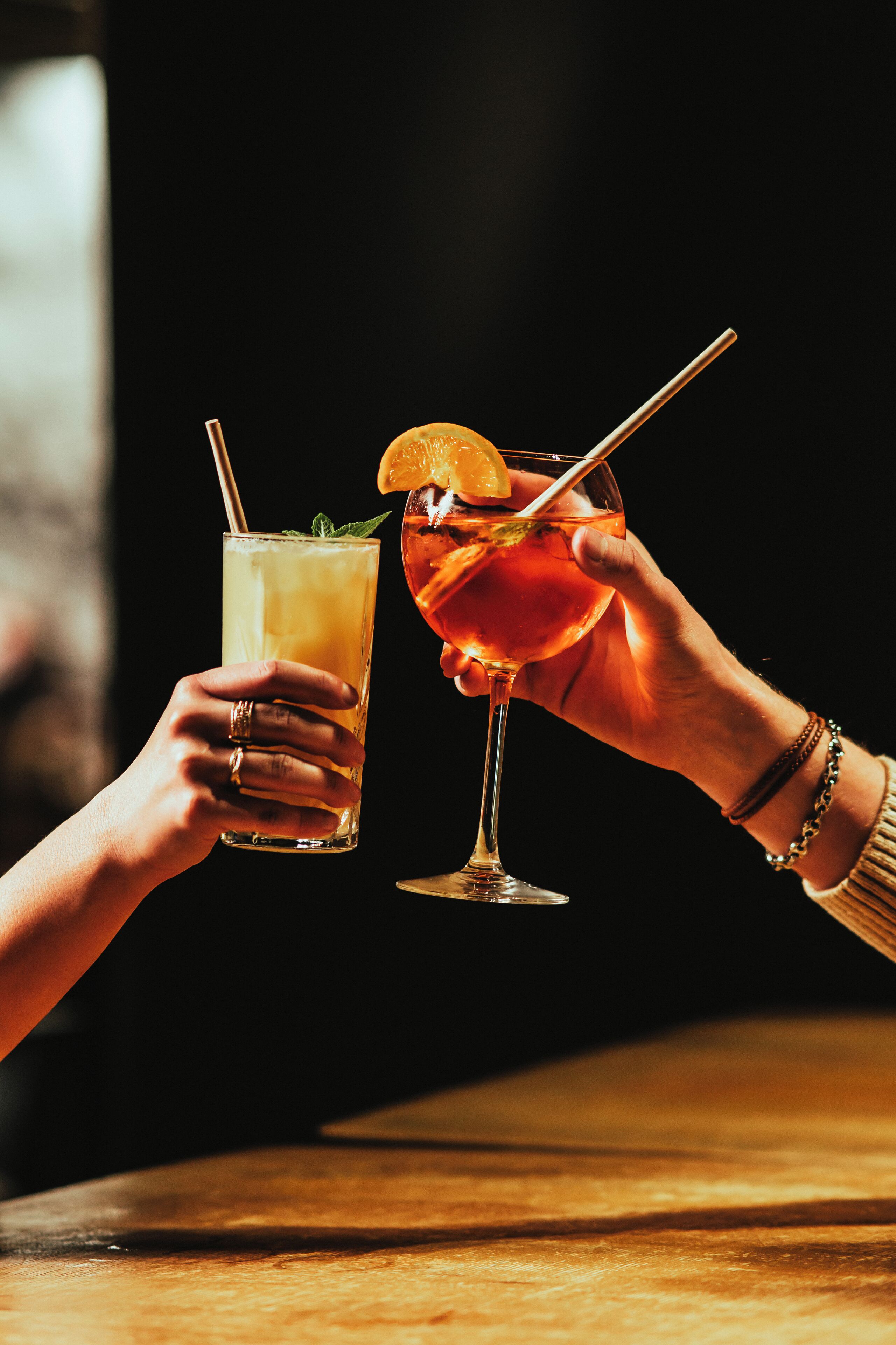 Two hands holding glasses of a beverage, one in a tall glass and one in a stemmed glass, with a dark background.