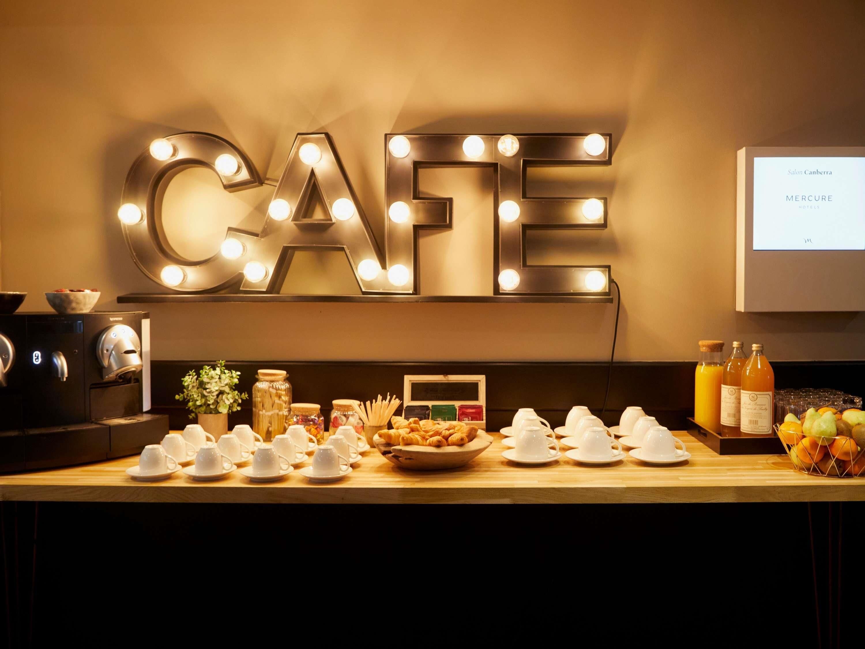 A cafe with illuminated letters spelling 'CAFE' on the wall, a coffee machine, and a variety of food and drink items on the counter.