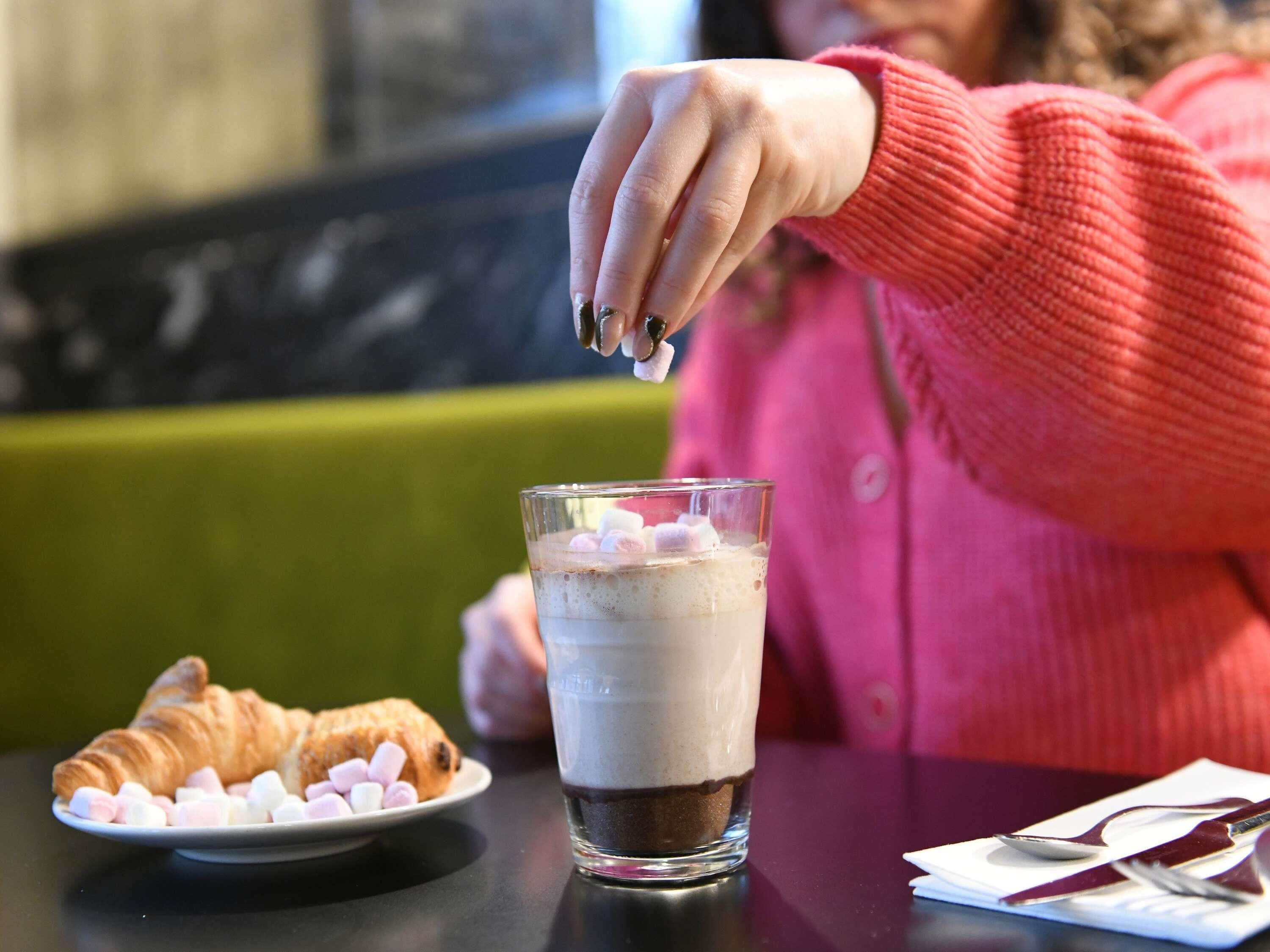 Eine Person sitzt an einem Tisch mit einem Glas mit einem schichtartigen Getränk, einem Teller mit Marshmallows und einem Croissant.