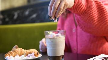 Eine Person sitzt an einem Tisch mit einem Glas mit einem schichtartigen Getränk, einem Teller mit Marshmallows und einem Croissant.