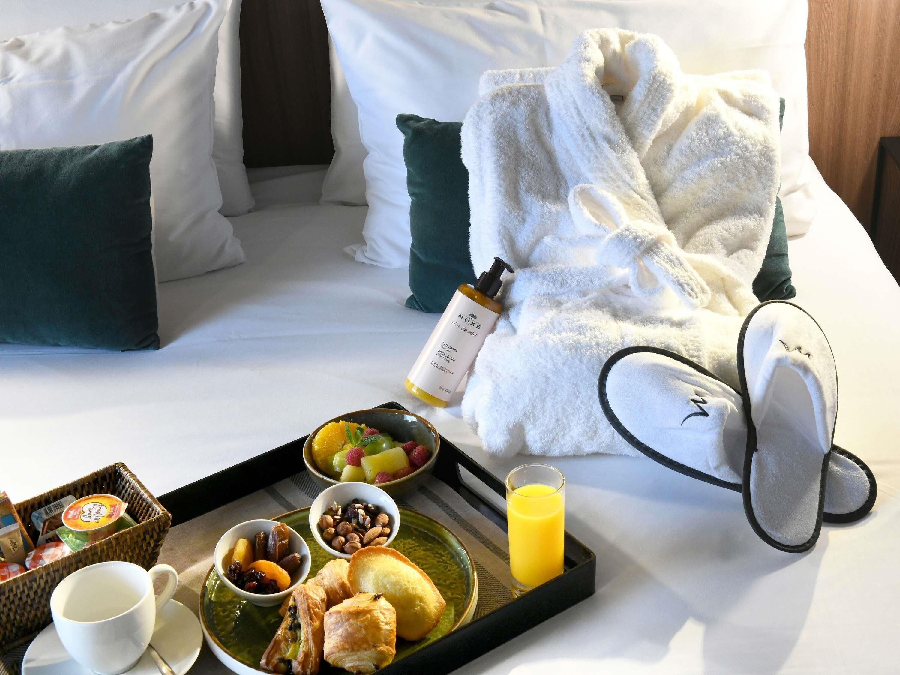 Un lit bien fait, avec des oreillers blancs et verts, un plateau de petit-déjeuner comprenant des fruits, des noix, des viennoiseries et un verre de jus d'orange.