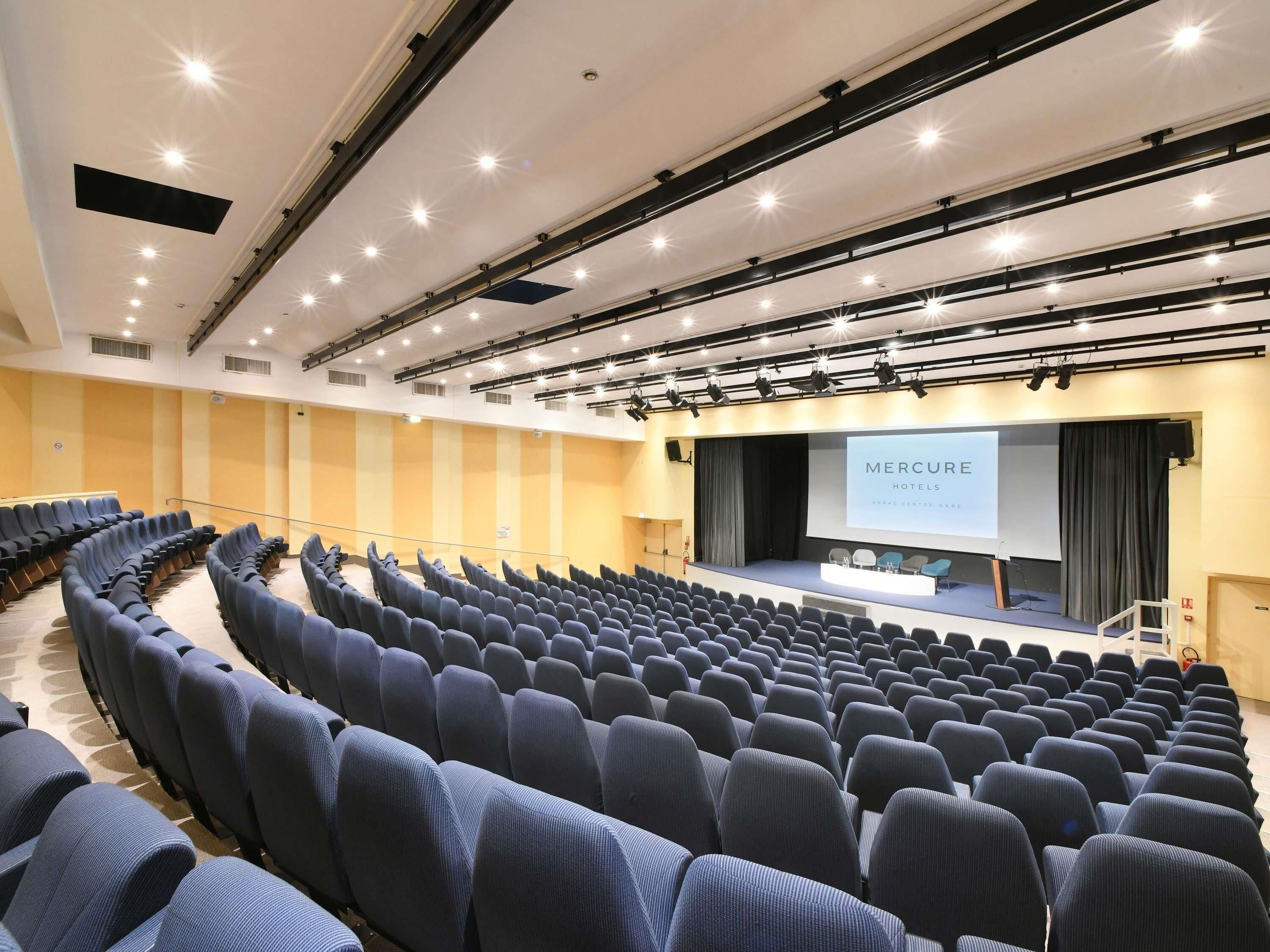 A conference room with rows of chairs, a stage with a screen displaying 'MERCURE HOTELS,' and a large window.