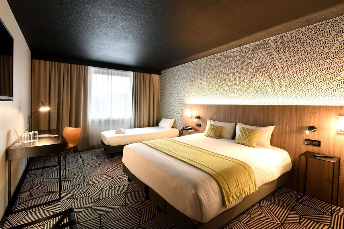 A hotel room with a large bed, a desk with a chair, a small table, and a patterned carpet.