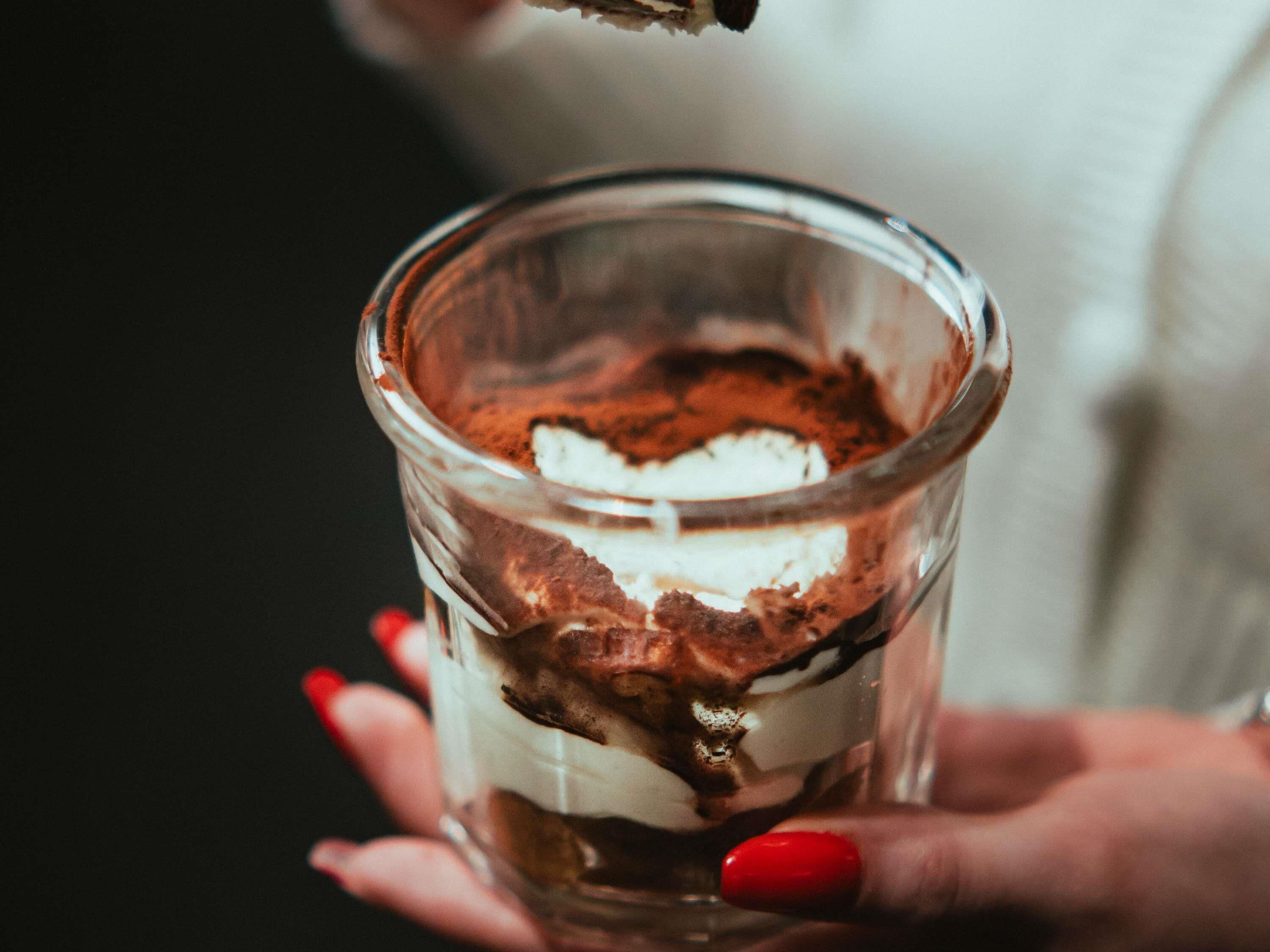A hand holding a layered dessert in a clear glass.