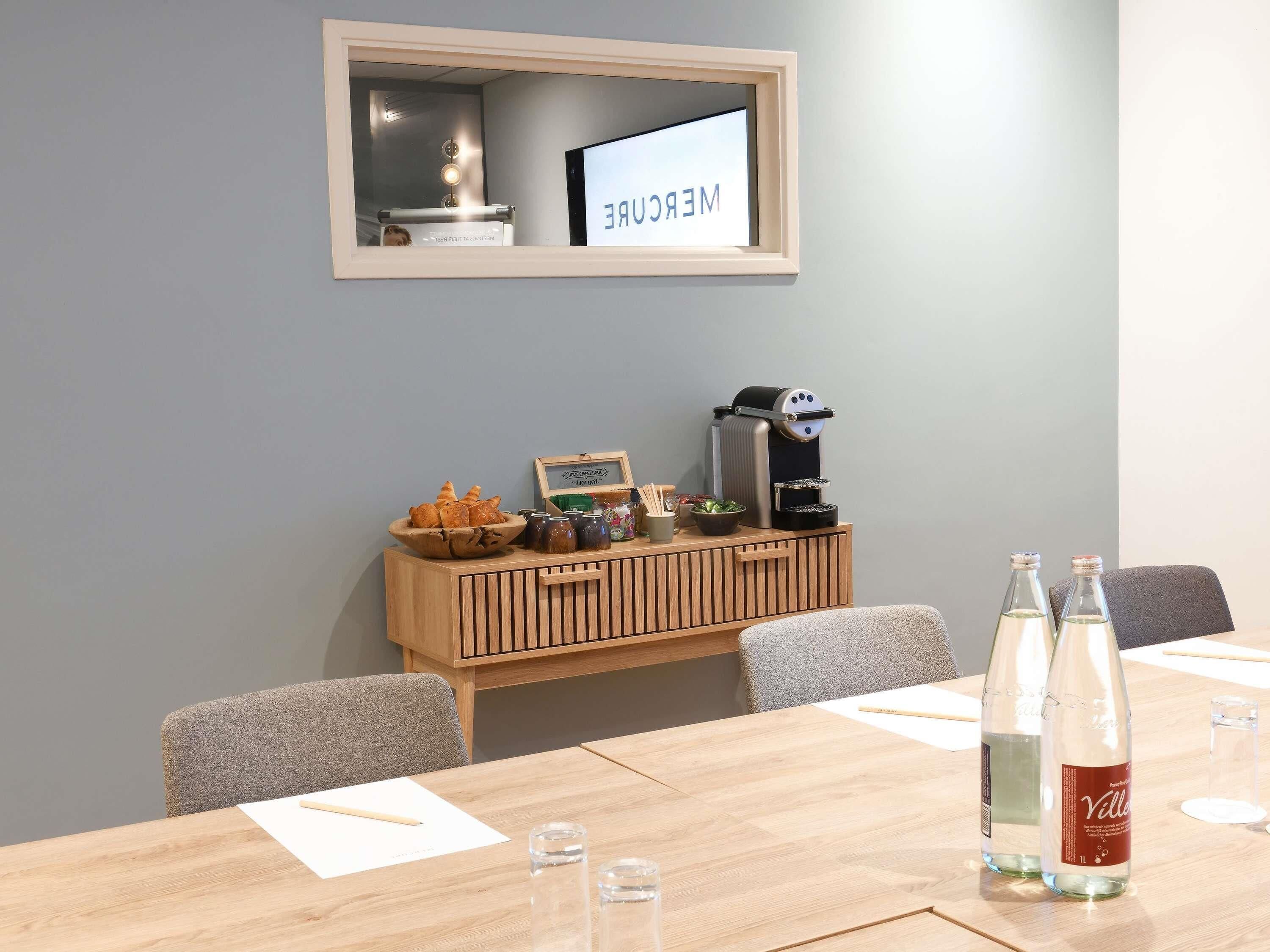 A conference room with a wooden table, chairs, a coffee machine, and a small display case with pastries.