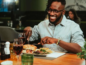 A person dining at a restaurant with a plate of food, a glass of red wine, and condiments on the table.