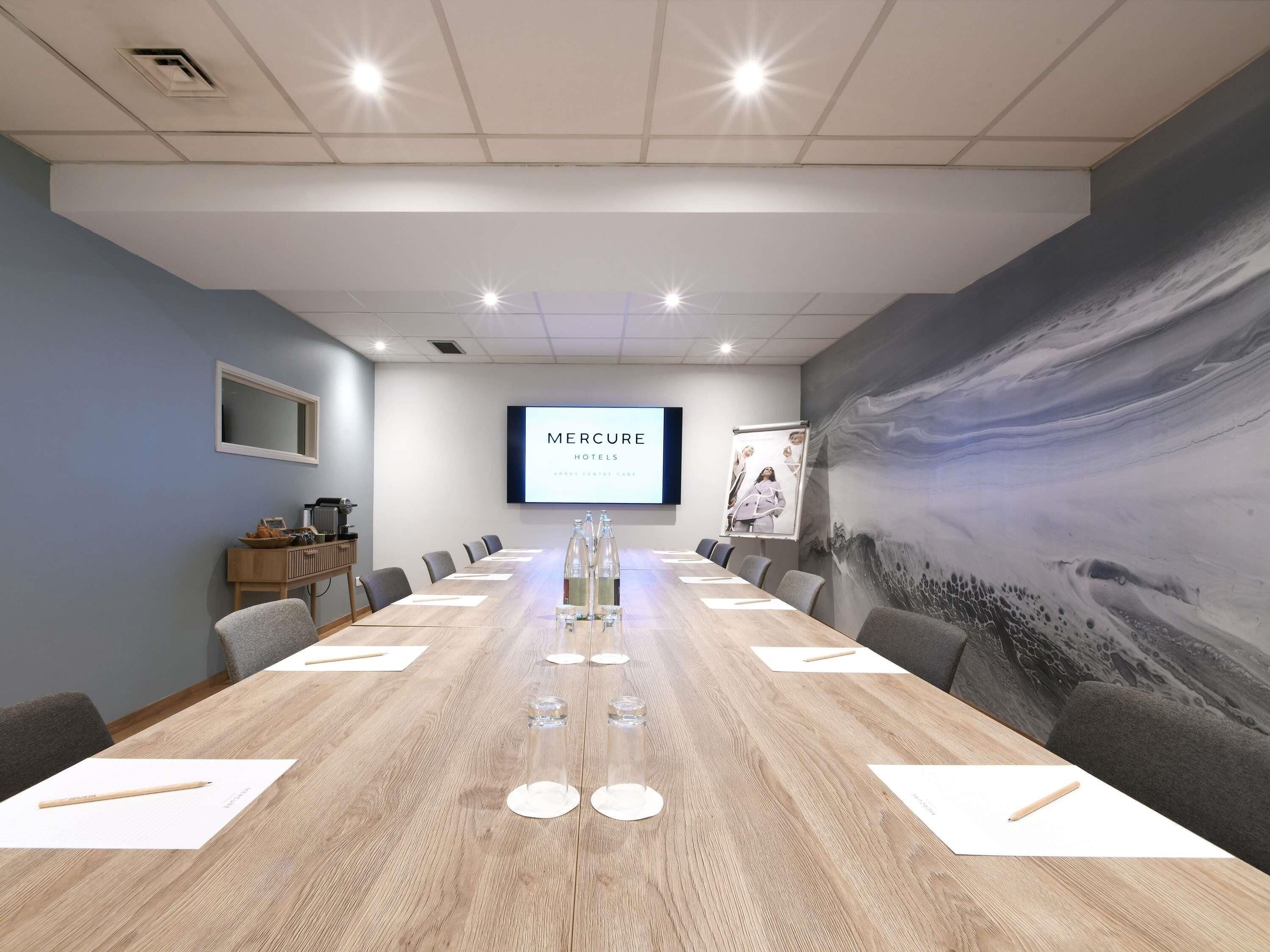 Ein Konferenzraum mit einem langen Tisch, Stühlen, einer Projektionsleinwand, die 'MERCURE HOTELS' anzeigt, und einem großen, abstrakten Wandbild.