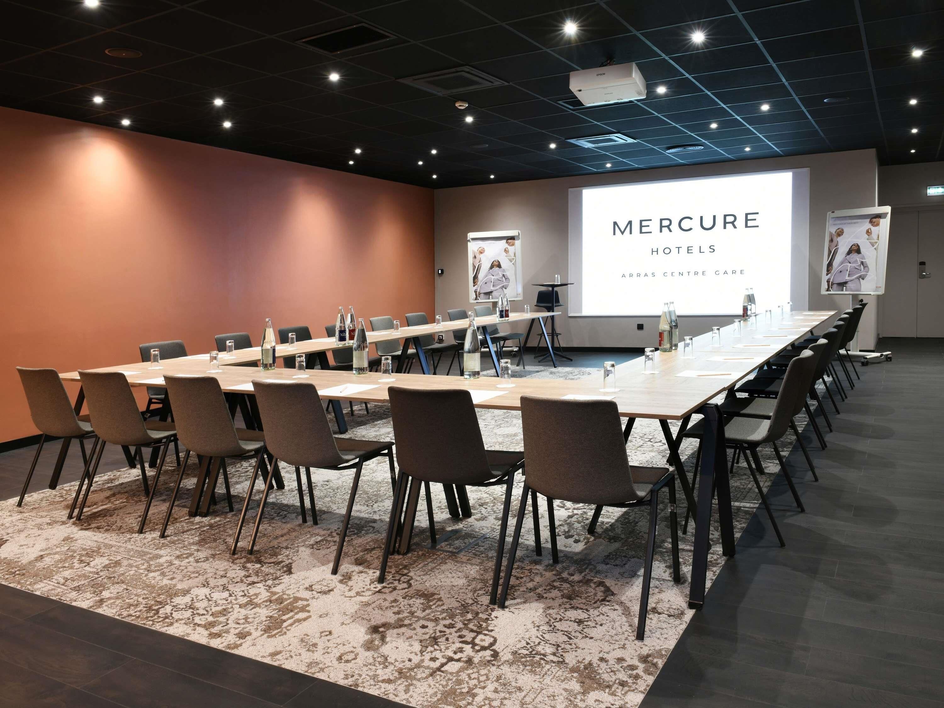 Un salón de conferencias con una larga mesa, sillas, una pantalla de proyector y una presentación titulada 'MERCURE HOTELS ARRAS CENTRE GARE'.