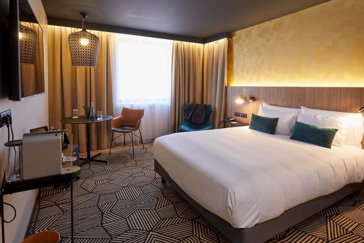 A hotel room with a large bed, a desk with a chair, a small table with a chair, and a large window with curtains.