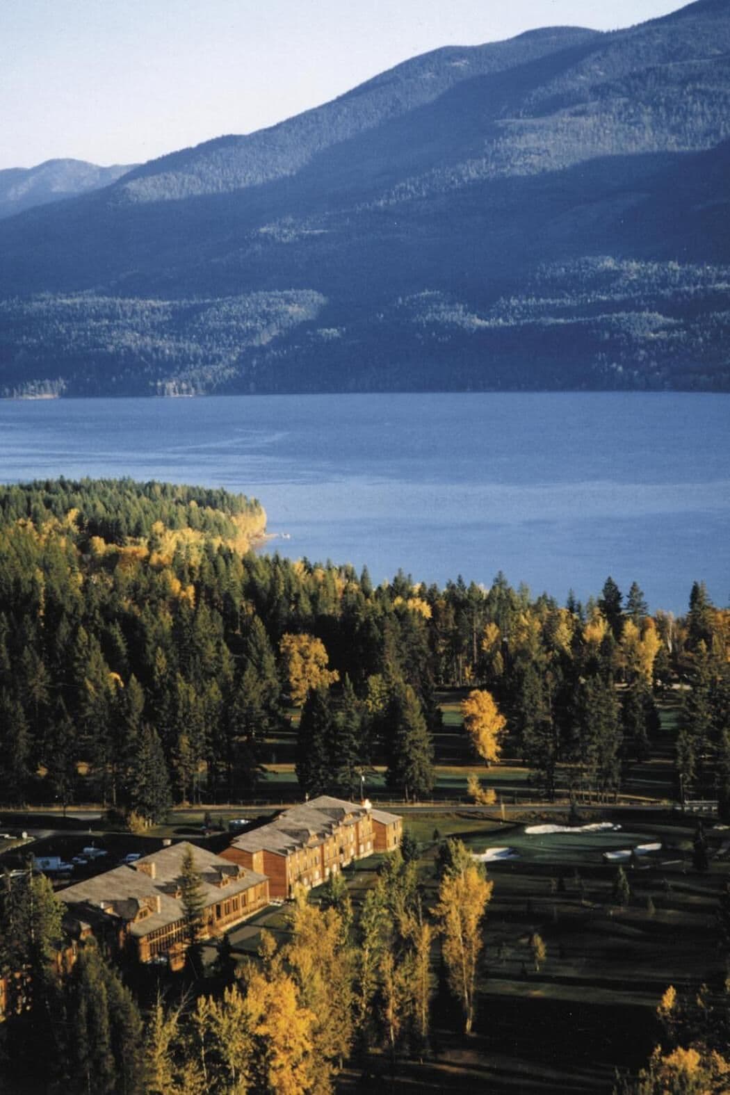 A large building with multiple sections, surrounded by dense forest and a body of water.
