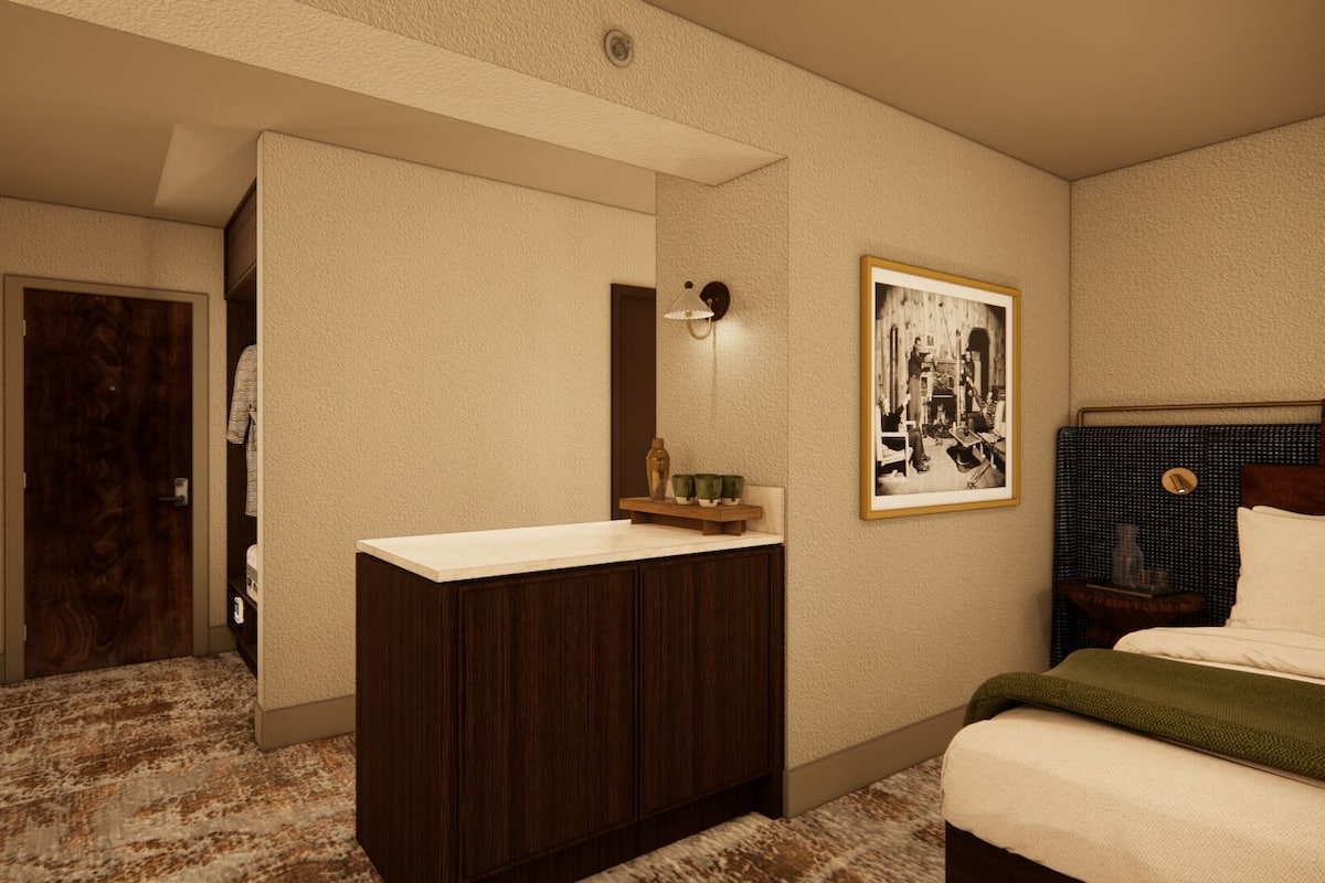 A hotel room with a wooden vanity, a bed with a green throw, a framed black and white photo on the wall, and a patterned carpet.