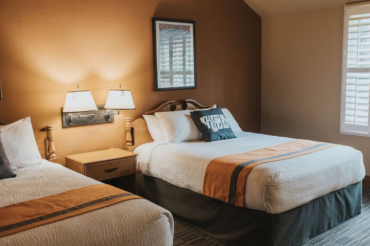 A hotel room with two beds, a nightstand, a lamp, a framed picture, and a window with blinds.