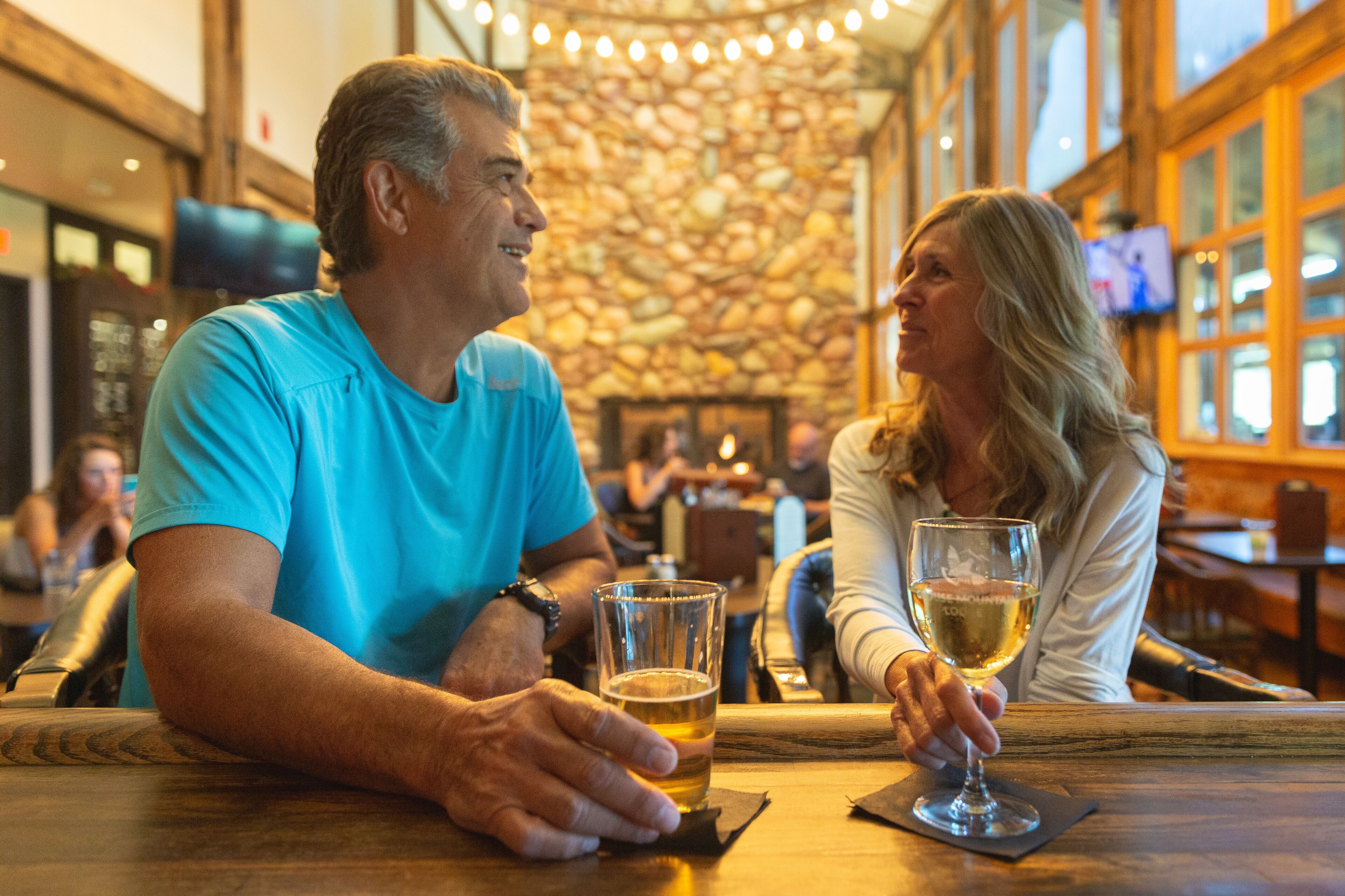 Un homme et une femme sont assis à une table en bois dans un restaurant. On aperçoit, en arrière-plan, un mur de pierre et des poutres en bois.