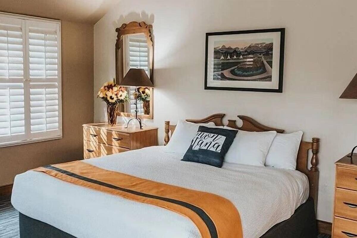 A neatly arranged hotel room with a large bed, a wooden dresser, a nightstand with a lamp, and a framed picture on the wall.