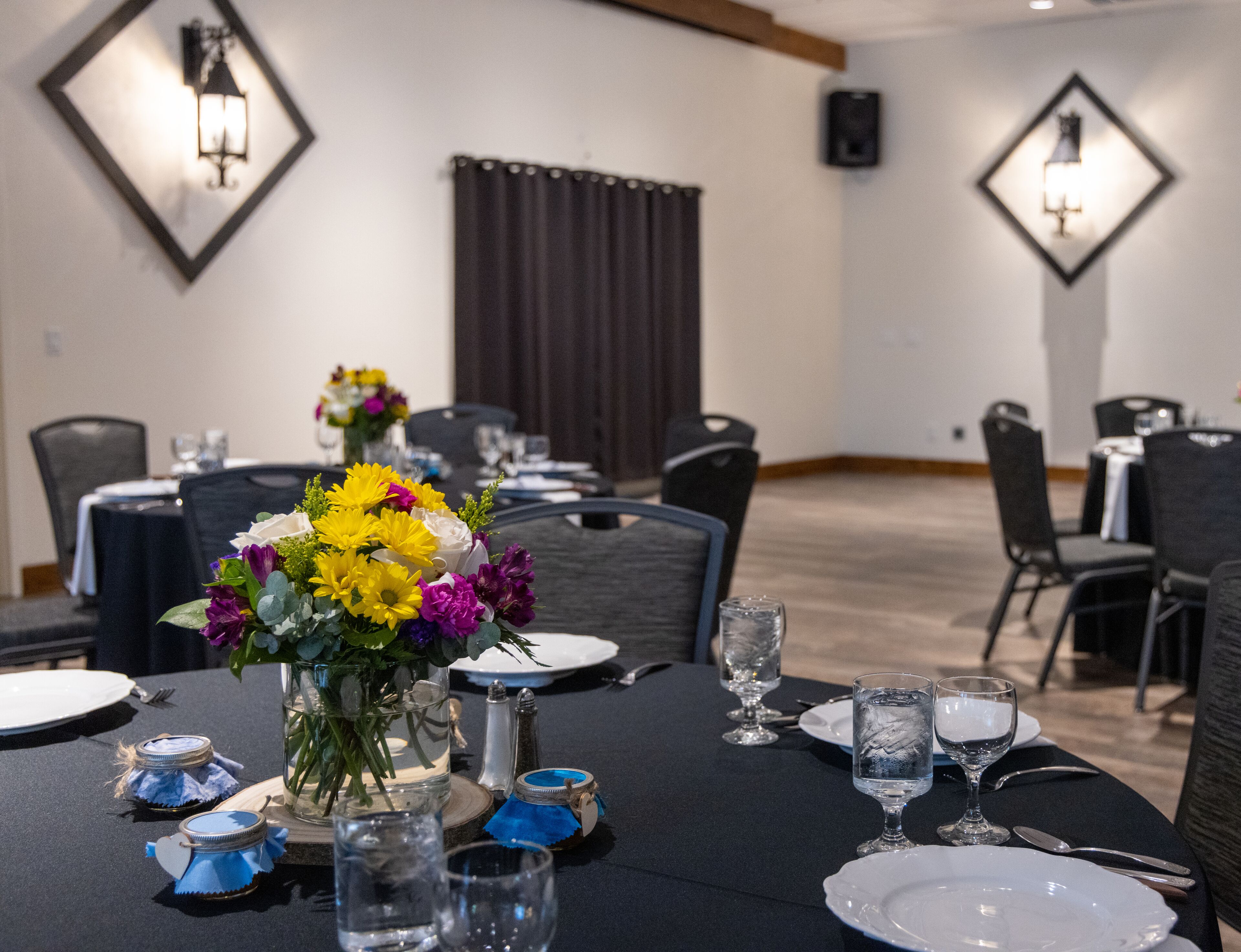 Uma sala de jantar com mesas postas para refeição, com centros de mesa florais, copos de vidro transparente e arranjos de mesa bem organizados.