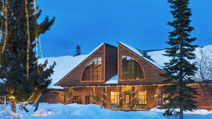 Una cabaña de troncos con ventanas amplias y techo cubierto de nieve, rodeada de nieve y decorada con luces.