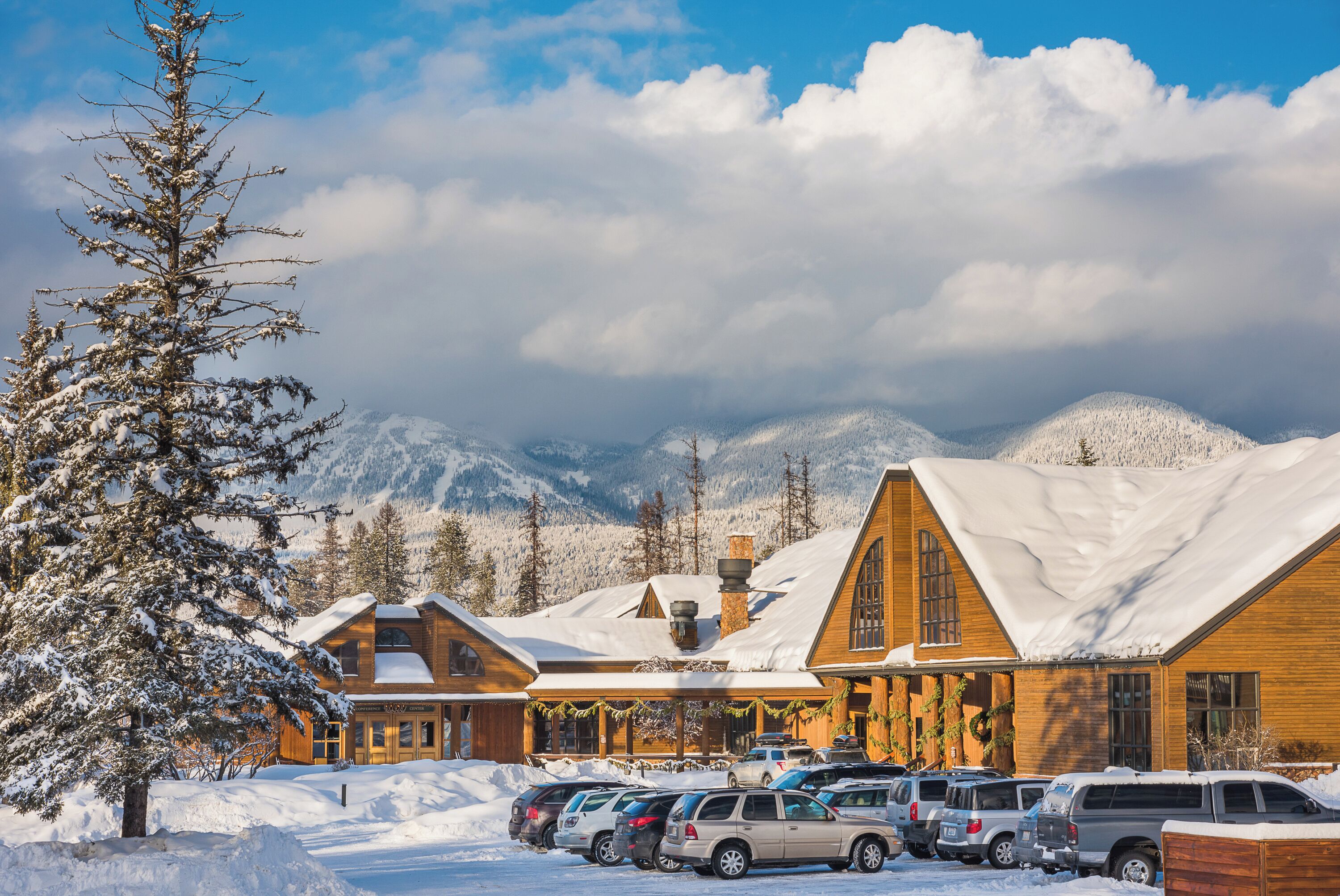 Un albergue con varias construcciones de madera, un estacionamiento con varios autos y montañas nevadas al fondo.