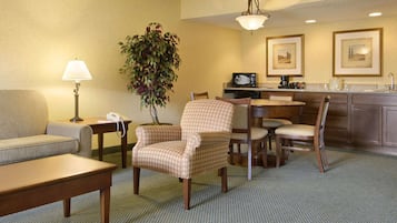 A hotel room with a sofa, armchair, dining table, chairs, a small kitchen area, and a plant.
