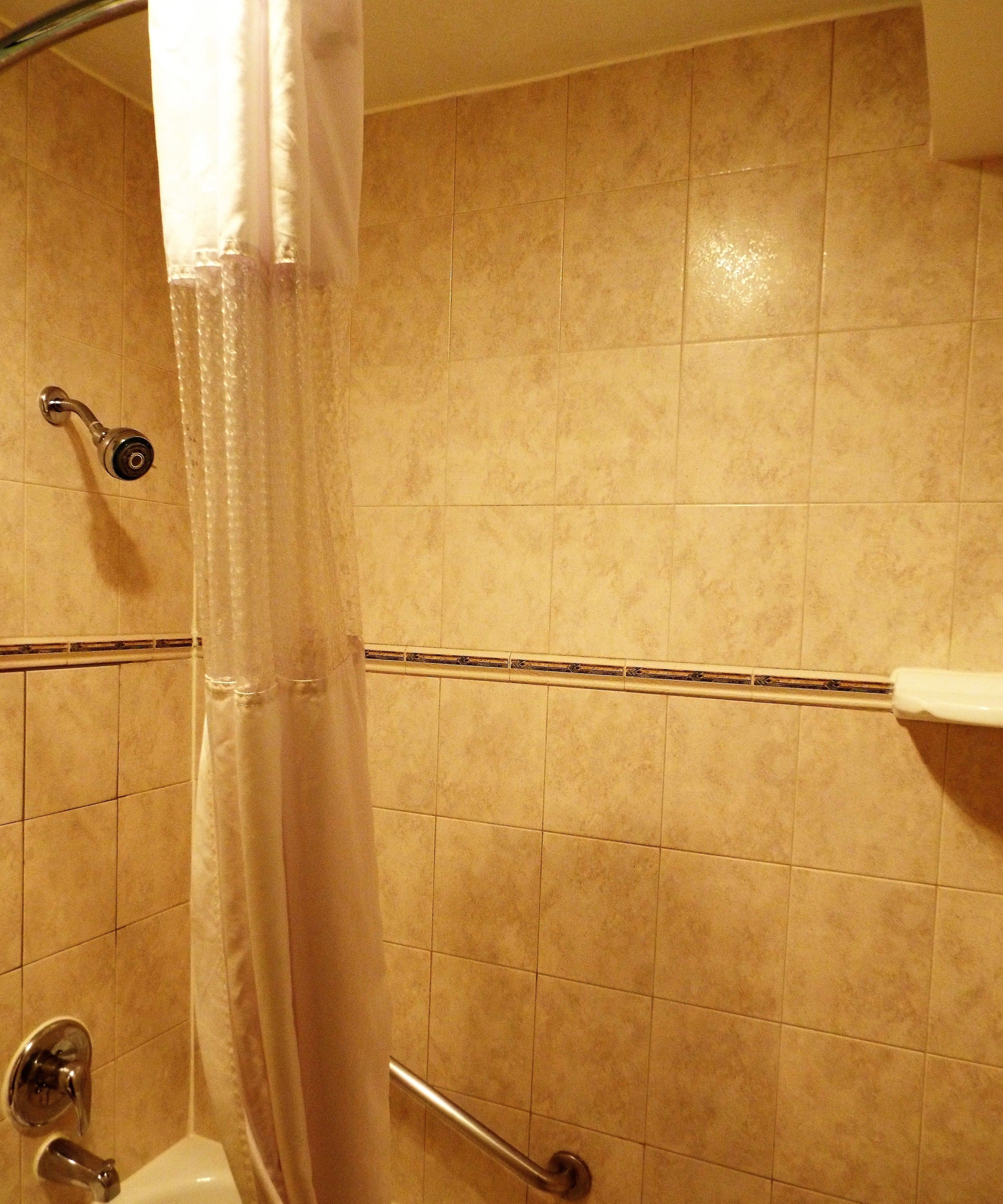 A bathroom with beige tiles, a shower curtain, a handheld showerhead, a grab bar, and a toilet.