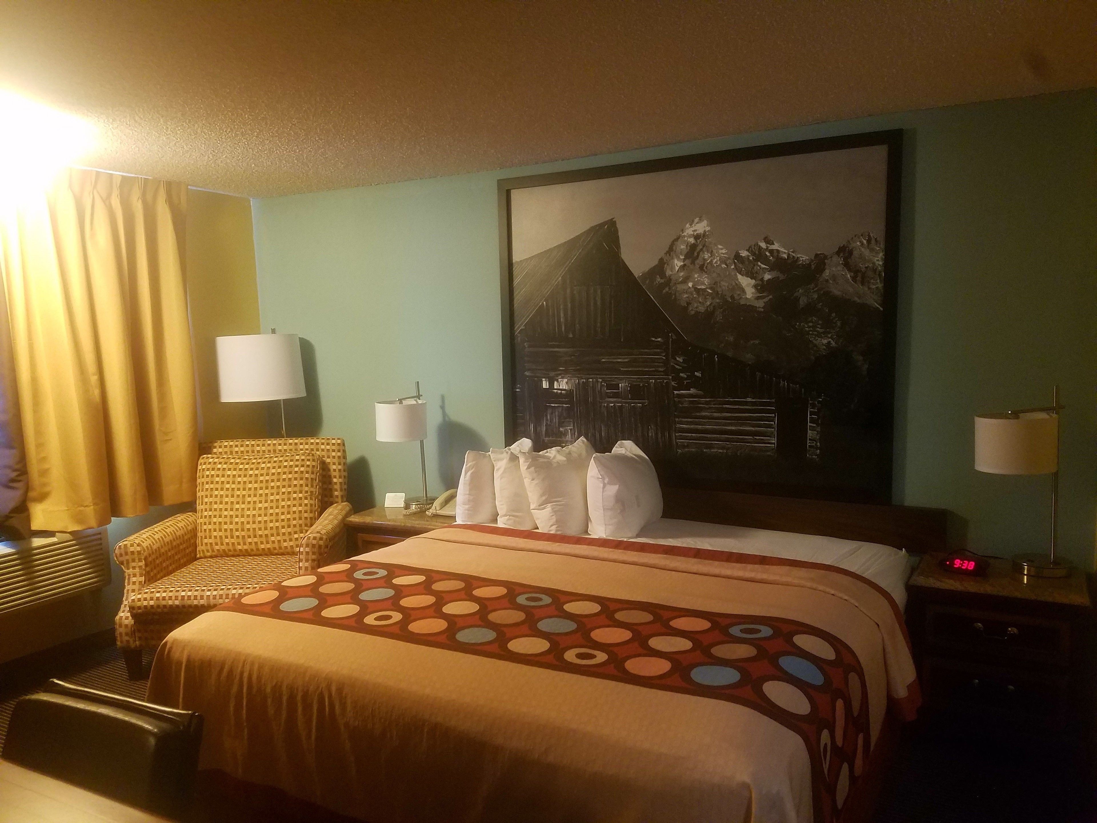 Una habitación de hotel con una cama, una silla, dos mesitas de noche con lámparas, un cuadro de montañas en la pared y un reloj.