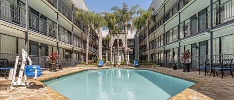 Una piscina con plataforma de baldosas, rodeada por un edificio con balcones y palmeras.