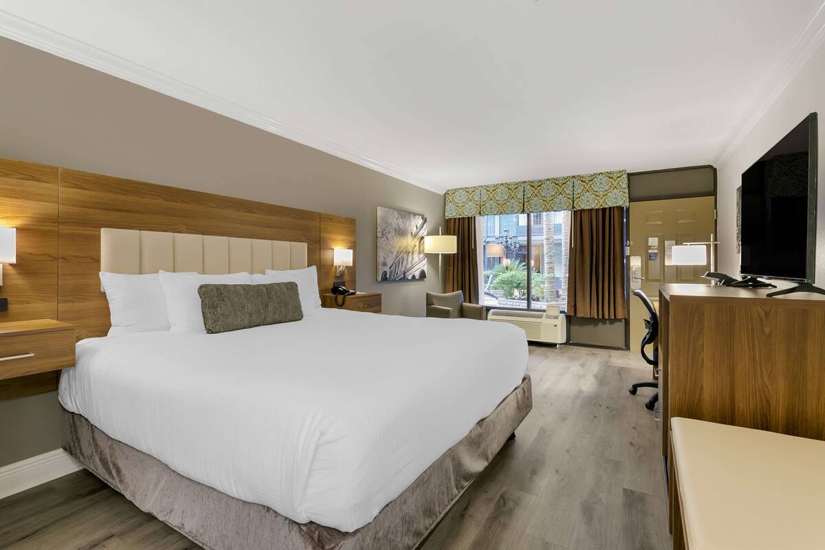 Habitación de hotel con una cama grande, mesitas de noche con lámparas, un escritorio, una silla, un televisor y una ventana con cortinas.