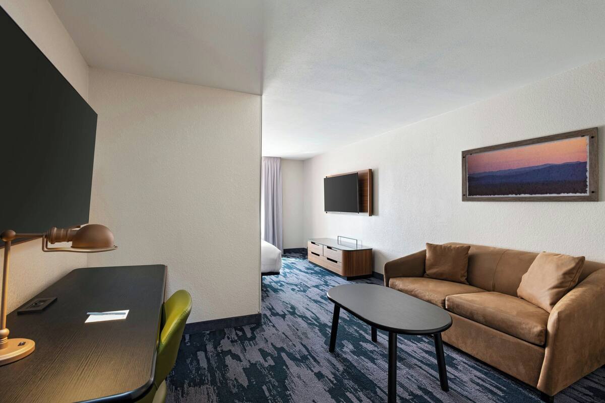 A hotel room with a bed, a sofa, a desk with a lamp, a TV, and a painting on the wall.