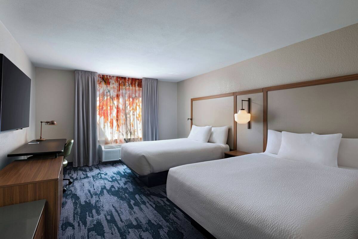 A hotel room with two beds, a desk, a flat-screen TV, and a large window with curtains.
