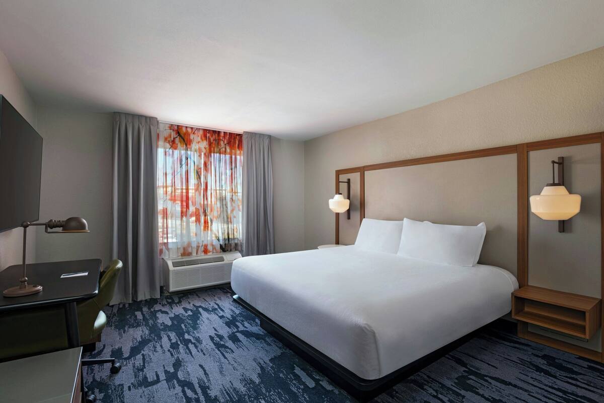 A hotel room with a large bed, a desk with a lamp, a window with curtains, and a wall-mounted light fixture.