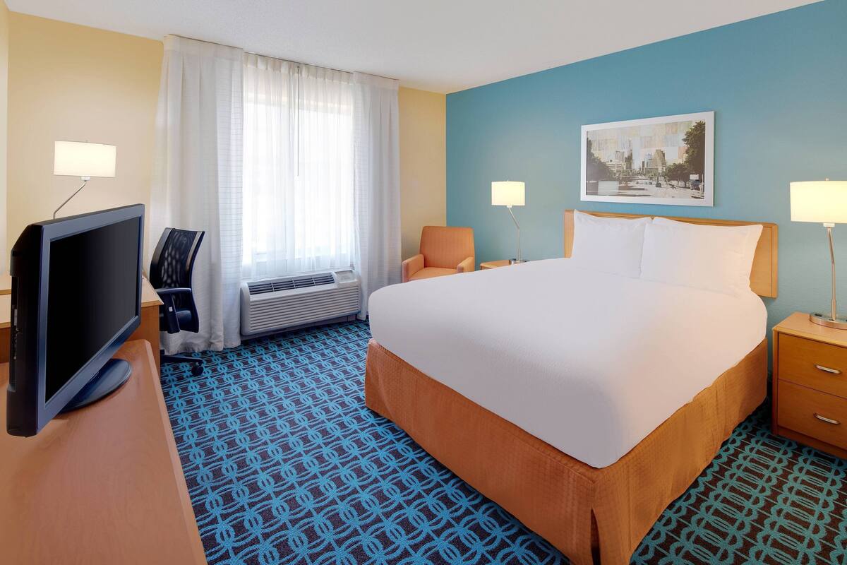A hotel room with a large bed, a desk with a television, a chair, a window with curtains, and a painting on the wall.