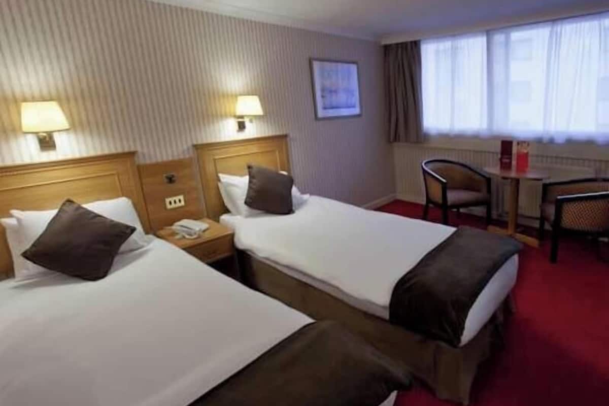 A hotel room with two beds, a small table, a chair, and a window with curtains.