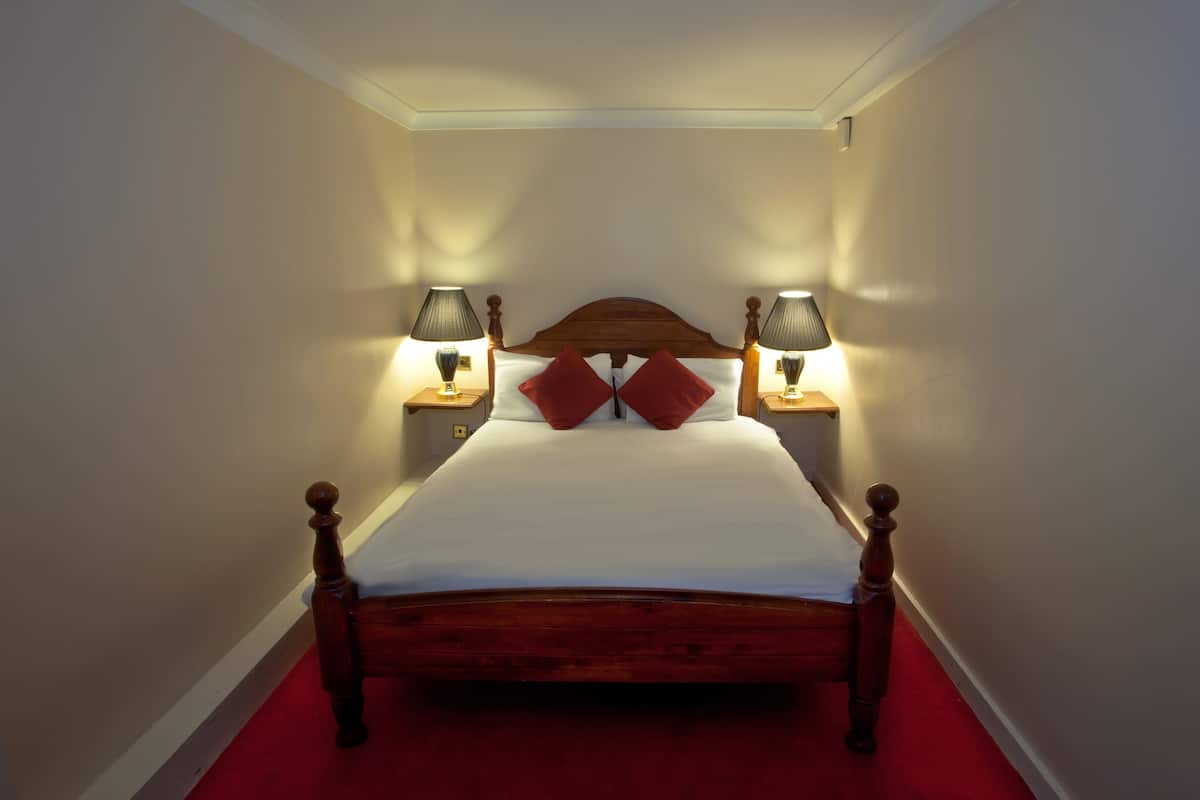 A neatly made bed with a wooden headboard, two bedside lamps, and a red carpet.