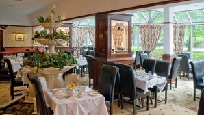 Un restaurante con mesas dispuestas para comer, una fuente decorativa como pieza central y ventanas grandes con cortinas.