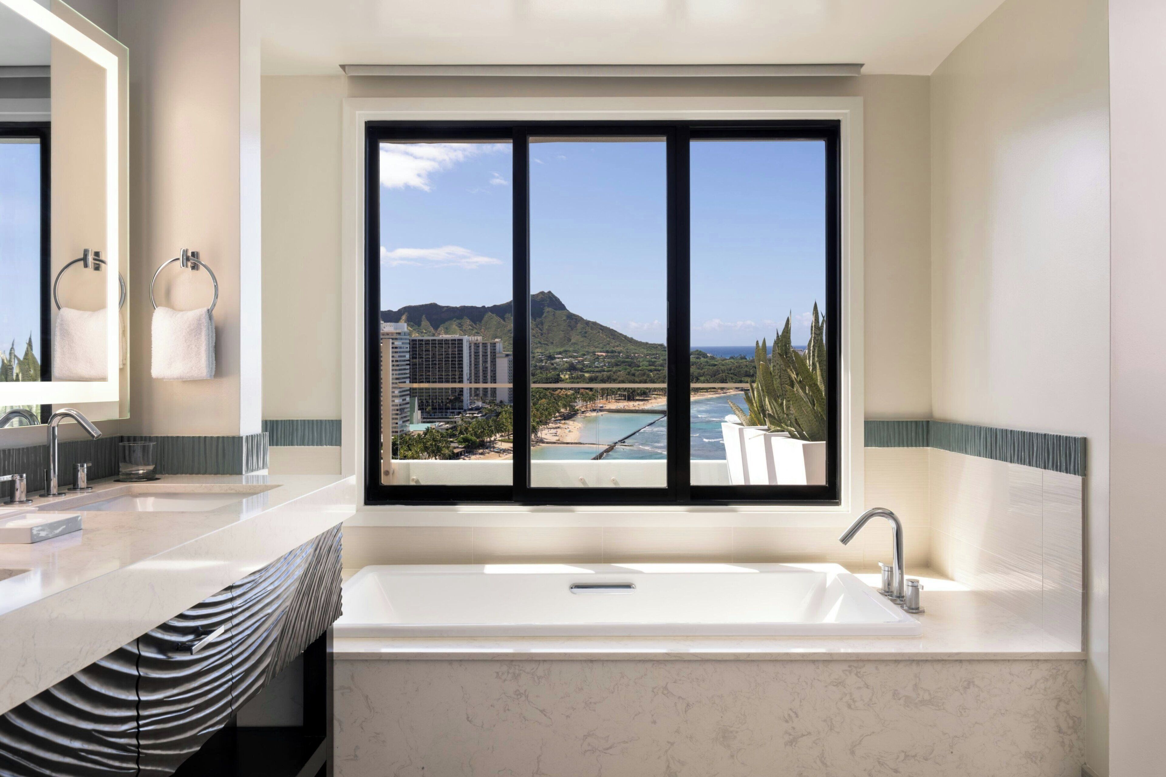 A modern bathroom with a large bathtub, dual sinks, and a window offering a view of a beach and buildings.