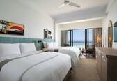 A hotel room with two beds, a ceiling fan, a large window with a view, and a beach-themed artwork on the wall.