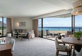 A modern hotel room with a balcony, a sofa set, a dining table, and a large window offering a view of the sea.