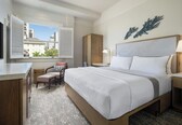 A hotel room with a bed, a desk, a chair, a window with blinds, and a view of buildings.