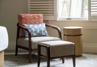 A chair with a patterned cushion and a matching ottoman, a wooden side table, and a window with blinds.