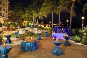 An outdoor event space with round tables covered in blue tablecloths, arranged on a paved area with palm trees and a view of a cityscape in the background.