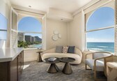 A modern living room with a sofa, two coffee tables, a chair, and large windows with a view of the ocean and buildings.